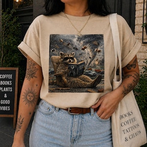 May include: Beige t-shirt featuring a raccoon reading a book while a tornado rages in the background. The raccoon is seated in a chair. The shirt also has the text "BOOKS COFFEE TATTOOS & CHAOS" on a tote bag.