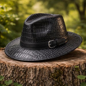 Może przedstawiać: Czarny kapelusz typu fedora o fakturze skóry krokodyla. Kapelusz posiada skórzany pasek ze srebrną klamrą. Kapelusz leży na pniu drzewa z rozmytym zielonym tłem.