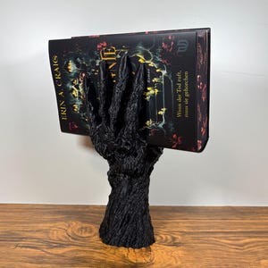 Könnte beinhalten: Ein schwarzer Buchhalter in Form eines Baumes, der ein Buch hält. Das Buch hat einen dunklen Einband mit floralen und Fantasy-Bildern. Der Buchhalter besteht aus einem strukturierten Material, und das Buch wird von einer handartigen Struktur gehalten.
