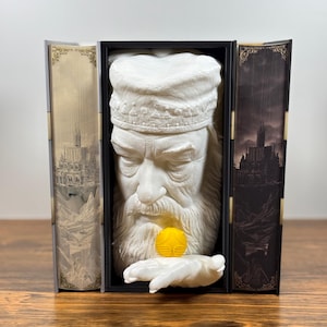 Könnte beinhalten: Eine weiße Büste eines bärtigen Mannes mit Hut, der eine goldene Kugel in einer weißen Hand hält. Die Büste ist in einem schwarzen Rahmen eingefasst, flankiert von Tafeln mit Schlossillustrationen. Der Hintergrund ist eine Holzoberfläche.