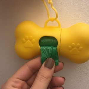 Könnte beinhalten: Gelber, knochenförmiger Hundekotbeutelspender mit einem teilweise herausgezogenen grünen Beutel. Der Spender hat Pfotenabdruckdetails und einen gelben Clip zur einfachen Befestigung.