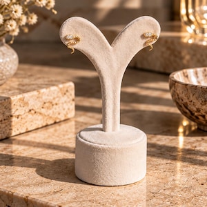 Può includere: Un paio di orecchini a lobo a forma di serpente dorato, con piccole pietre trasparenti incastonate nella testa. Gli orecchini sono esposti su un supporto per gioielli in velluto beige a forma di Y, che mette in risalto il design curvo.