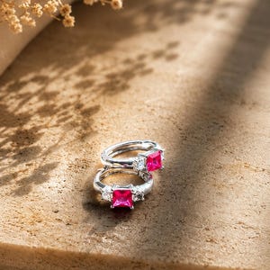 May include: A pair of silver hoop earrings, each featuring a prominent square-cut vibrant pink gemstone at the center, flanked by two smaller clear round stones on either side, set within a polished metallic band with a secure hinged closure mechanism.