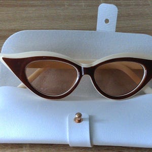 May include: Cat-eye sunglasses with a cream-colored frame and brown lenses rest in a white case. The sunglasses have a dark brown accent on the top frame. The case is closed with a gold-colored button.