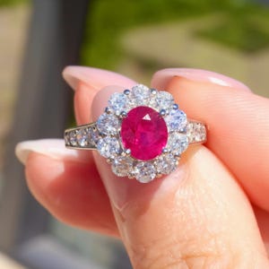 May include: A silver ring with a central oval ruby, encircled by a halo of smaller diamonds. The band is also set with diamonds. The ring's gemstones are set off against a blurred green backdrop.
