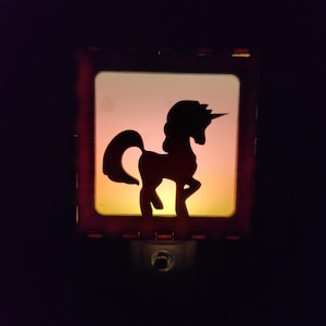 Pode incluir: Uma luz noturna quadrada com a silhueta preta de um unicórnio contra um fundo gradiente brilhante de tons quentes, que transita de um amarelo suave na base para um laranja claro no topo, dentro de uma moldura escura e sólida.