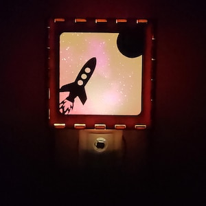 May include: A square night light featuring a black silhouette of a rocket ship launching into a pink and white nebula background with a dark circular planet in the upper right corner, all enclosed within a dark, notched frame that glows from within.