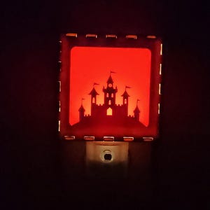 May include: A square night light featuring a glowing red background with a black silhouette of a castle with multiple towers and flags. The frame is dark with small rectangular cutouts along the edges, and a standard plug base is visible below.