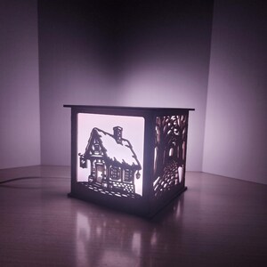 May include: A square wooden lamp with laser-cut panels featuring a cottage with a snow-covered roof on one side and a cobblestone path lined with trees on the other. The lamp emits a soft white glow from within, highlighting the intricate cutouts.