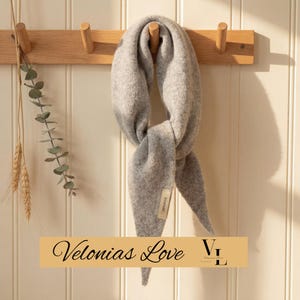 May include: A pale grey wool scarf hangs from a wooden peg rail. The scarf, with a soft texture, has a label reading "100% wool." The rail is fixed to a white panelled wall, with decorative sprigs of greenery and wheat stalks to the left.