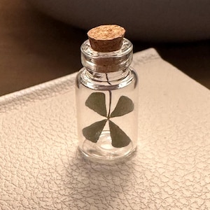 May include: A miniature glass bottle with a cork, holding a four-leaf clover. The bottle sits on a textured, cream-coloured surface. The clover is green, and the bottle is small. The background includes a grey object and a wooden surface.