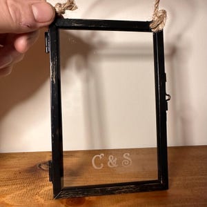 Op de afbeelding: Zwart metalen en glazen fotolijst met een verweerde afwerking, opgehangen aan een natuurlijk touw. De lijst heeft de geëtste initialen "C & S" op het glas. De lijst wordt door een hand vastgehouden.