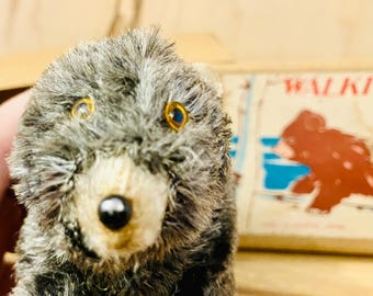 Pode incluir: Uma pequena figura de urso cinzento e felpudo com olhos cor de âmbar e nariz preto. Atrás do urso encontra-se uma caixa retangular com o texto WALKIE e uma ilustração gráfica de um urso castanho num cenário azul e branco.