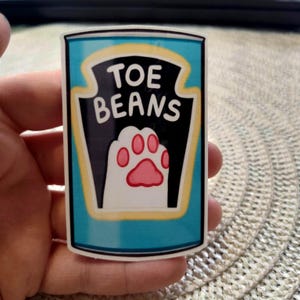 May include: A rectangular sticker with a blue background and a black and white label. The label reads "TOE BEANS" and features a pink paw print. The sticker has a yellow border.