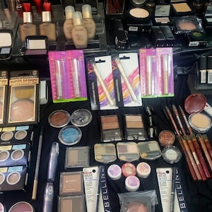 May include: A collection of various makeup products, including eyeshadow palettes, lipsticks, foundation bottles, and brushes. The products are arranged on a black surface, showcasing a variety of colors and textures. Several items are packaged in clear plastic.