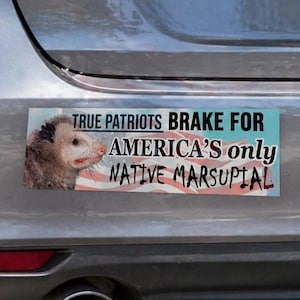 May include: A rectangular bumper sticker with the text "TRUE PATRIOTS BRAKE FOR AMERICA'S ONLY NATIVE MARSUPIAL" and an image of an opossum. The background features a faded American flag design.