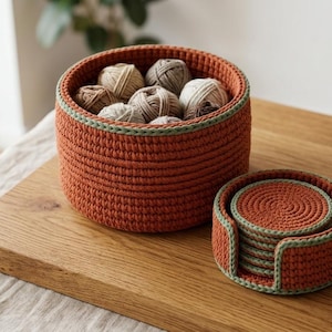 Op de afbeelding: Een ronde, roestoranje gehaakte mand gevuld met verschillende kleine bolletjes garen in neutrale tinten, vergezeld door een bijpassende ronde gehaakte houder met een stapel ronde roestoranje onderzetters met een saliegroene rand.