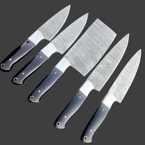 Op de afbeelding: Een set van vijf keukenmessen met patronen stalen messen en zwarte handvatten. De messen hebben een unieke, getextureerde uitstraling. De handvatten zijn zwart met een zilveren bolster en een decoratieve zilveren klinknagel. Op een donkergrijze achtergrond.