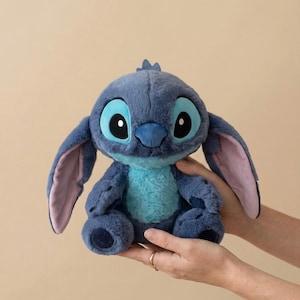 May include: A blue Stitch plush toy, featuring large black and white eyes, a blue nose, and large ears with pink insides. The toy is from the Disney film Lilo & Stitch. The plush is held in two hands.