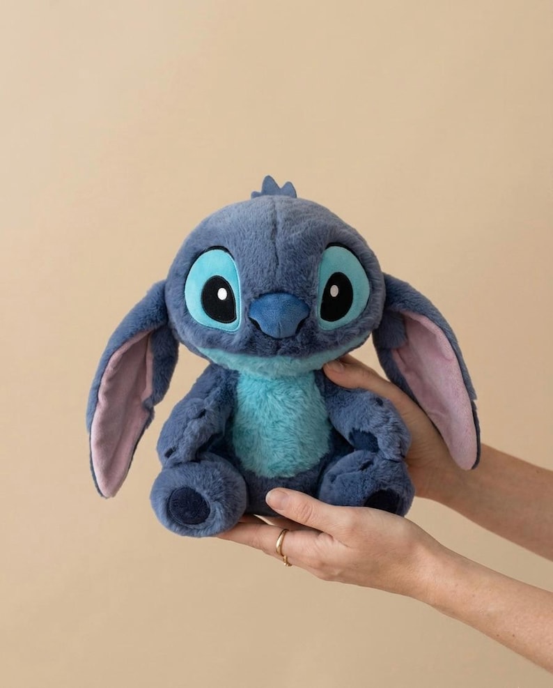 May include: A blue Stitch plush toy, featuring large black and white eyes, a blue nose, and large ears with pink insides. The toy is from the Disney film Lilo & Stitch. The plush is held in two hands.