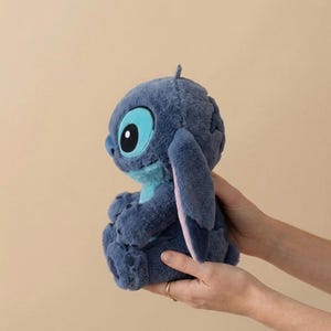 May include: A blue plush toy of the character Stitch from Lilo & Stitch. The furry creature has large ears and big eyes. The toy is held in two hands against a neutral background. The toy is a popular character from the Disney film.