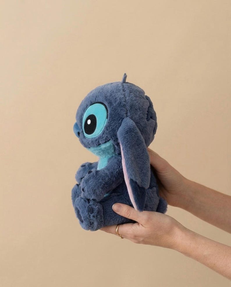 May include: A blue plush toy of the character Stitch from Lilo & Stitch. The furry creature has large ears and big eyes. The toy is held in two hands against a neutral background. The toy is a popular character from the Disney film.