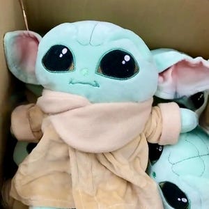 May include: Plush toy of a character with large black eyes, green skin, and large ears. The toy is wearing a tan coat and a beige scarf. Several of the toys are visible inside a cardboard box.