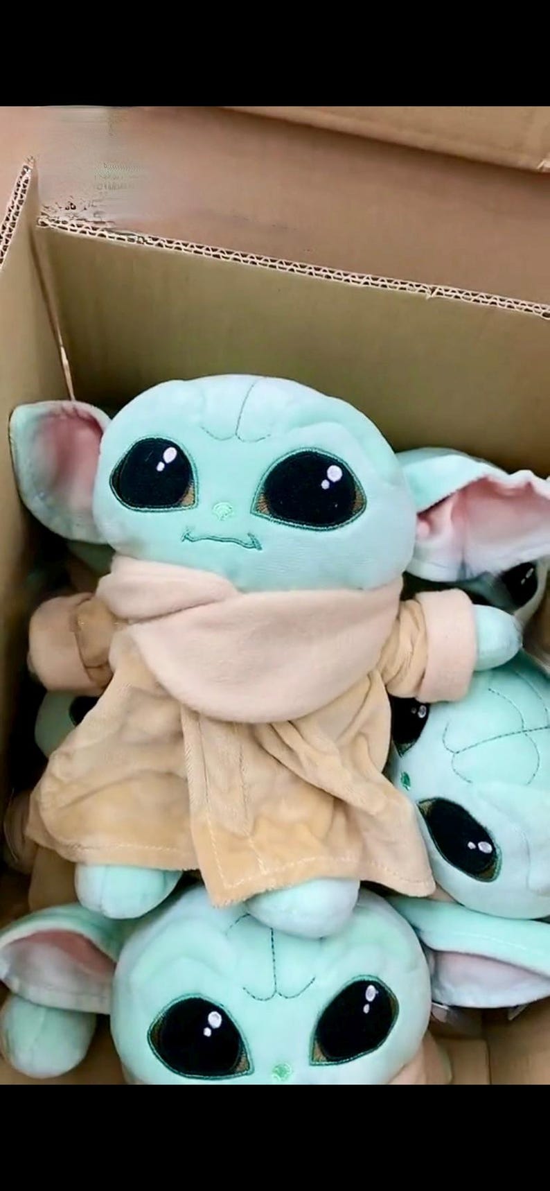 May include: Plush toy of a character with large black eyes, green skin, and large ears. The toy is wearing a tan coat and a beige scarf. Several of the toys are visible inside a cardboard box.