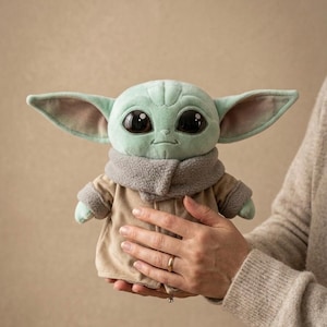 May include: A plush toy of a character with large black eyes, green skin, and large ears. The character wears a tan coat with a gray collar. The toy is held in front of a neutral background.