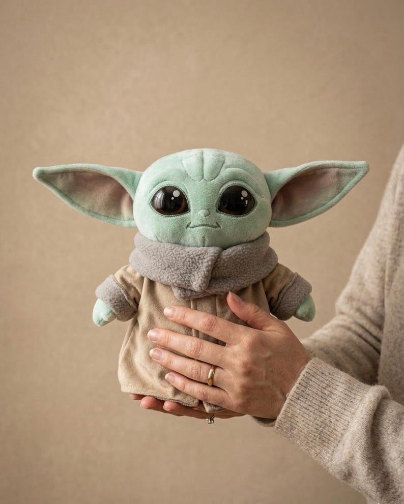 May include: A plush toy of a character with large black eyes, green skin, and large ears. The character wears a tan coat with a gray collar. The toy is held in front of a neutral background.