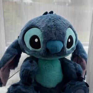 May include: A blue plush toy of the character Stitch, with large ears and expressive eyes. The toy has a turquoise belly and is sitting down. The toy is from the animated film Lilo & Stitch.