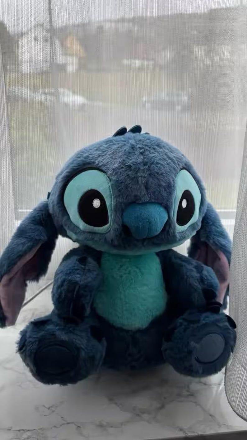 May include: A blue plush toy of the character Stitch, with large ears and expressive eyes. The toy has a turquoise belly and is sitting down. The toy is from the animated film Lilo & Stitch.