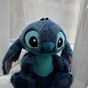 May include: A blue Stitch plush toy, featuring large eyes, ears, and a turquoise belly. The soft toy is positioned on a windowsill. The character is from the animated film Lilo & Stitch.