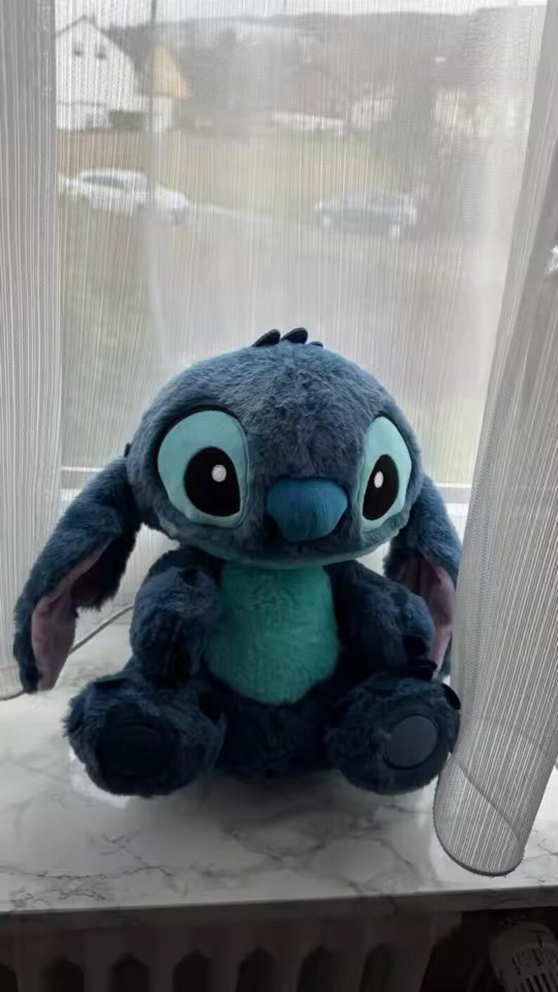 May include: A blue Stitch plush toy, featuring large eyes, ears, and a turquoise belly. The soft toy is positioned on a windowsill. The character is from the animated film Lilo & Stitch.