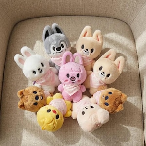 Peut inclure: Une collection de peluches en forme d'animaux, dont des lapins, des ours et un cochon. Les jouets sont dans des tons de blanc, rose, beige, gris et marron, certains avec des bandanas roses. Les jouets sont disposés sur une surface de couleur neutre.