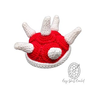 Puede incluir: Una concha de Koopa tejida a ganchillo en rojo y blanco. La concha presenta un cuerpo rojo con detalles y púas blancas. Este artículo de novedad es probablemente un accesorio de disfraz o una pieza decorativa. Hecho por Cozy Shell Crochet.
