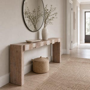 May include: A wooden console table with a decorative design, a round mirror, and a woven basket. A vase with greenery and a decorative bowl are on the table. A neutral-toned rug is on the floor.
