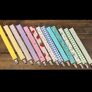 May include: A collection of twelve fabric pacifier clips arranged in a row, each featuring a metal clasp at one end and a loop at the other. The straps display various patterns including solid yellow, stripes, polka dots, gingham, strawberries, floral prints, and checkered designs in multiple colors.