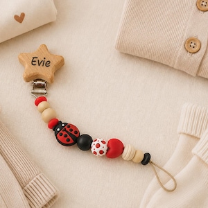 Könnte beinhalten: Ein Schnullerhalter aus Holz in Sternform mit dem eingravierten Namen "Evie", verziert mit einer Marienkäferperle und anderen bunten Perlen. Um den Halter herum befinden sich Babykleidung in neutralen Farbtönen, darunter eine Strickjacke, ein Body und Socken.