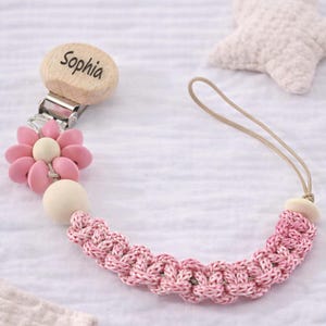May include: A pink and white pacifier clip with the name "Sophia" engraved on a wooden clip. The clip features a pink flower and a braided pink and white cord. A beige star-shaped item is in the background.