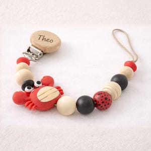 May include: A baby's dummy clip with a wooden clip engraved with the name "Theo". The clip features a red crab, wooden, black, and red beads. A beige string is attached to the end.