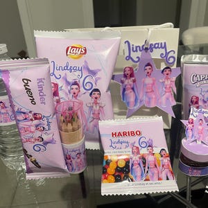 Może przedstawiać: Zestaw upominków i przekąsek na przyjęcie, w tym butelka wody, batonik Kinder Bueno, chipsy Lay's, cukierki Haribo, napój Capri Sun i mały pojemnik z kredkami. Wszystkie przedmioty mają fioletowo-biały motyw z imieniem "Lindsay".