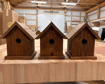 Puede incluir: Tres casas para pájaros de madera con tinte marrón oscuro. Cada casa para pájaros tiene un techo triangular, un orificio de entrada circular y una pequeña base cuadrada. Un gancho dorado está unido a la parte superior de cada casa.