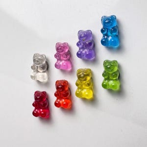 May include: A selection of colourful gummy bear sweets in shades of red, orange, yellow, green, blue, purple, and clear. The gummy bears are displayed on a white surface, highlighting their translucent quality.