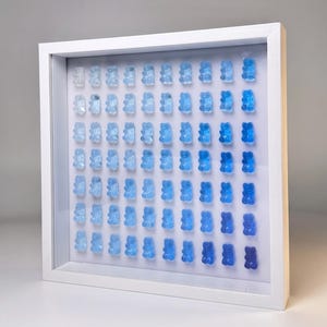 Peut inclure: Une œuvre d'art encadrée présentant une grille de bonbons oursons bleus. Les oursons changent de couleur, passant du bleu clair au bleu foncé, créant un effet de dégradé. L'œuvre est encadrée dans un cadre blanc.