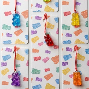 Könnte beinhalten: Eine Sammlung bunter Gummibärchen-Anhänger an roten Kordeln, präsentiert auf gemusterten Karten. Die Gummibärchen sind in verschiedenen Farben erhältlich, darunter Blau, Grün, Gelb, Rot, Orange und Lila. Die Karten haben einen weißen Hintergrund mit einem sich wiederholenden Muster von Gummibärchen.