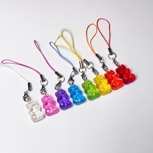 May include: A collection of eight translucent gummy bear charms in white, pink, purple, blue, green, yellow, orange, and red. Each bear is attached to a silver-toned metal lobster clasp and a thin, coloured nylon cord loop for hanging purposes.