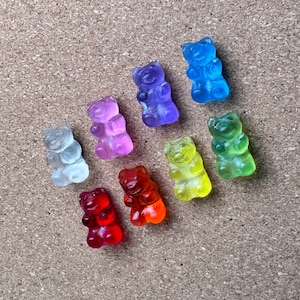 May include: An assortment of colorful gummy bear candies in various colors, including red, orange, yellow, green, blue, purple, and clear. The gummy bears are arranged on a textured, light brown surface.