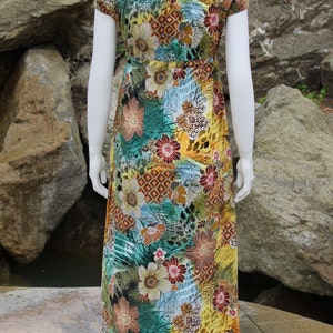 May include: A long, flowing maxi dress with a colorful floral print. The dress has a scoop neck, short sleeves, and a fitted bodice. The print features a variety of flowers, leaves, and geometric patterns in shades of yellow, green, blue, brown, and red.