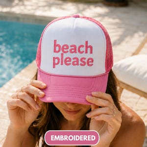 May include: A pink and white trucker hat featuring a white front panel with the text beach please embroidered in pink. The hat has a pink brim and pink mesh side and back panels. The design includes a structured front and an adjustable snapback.